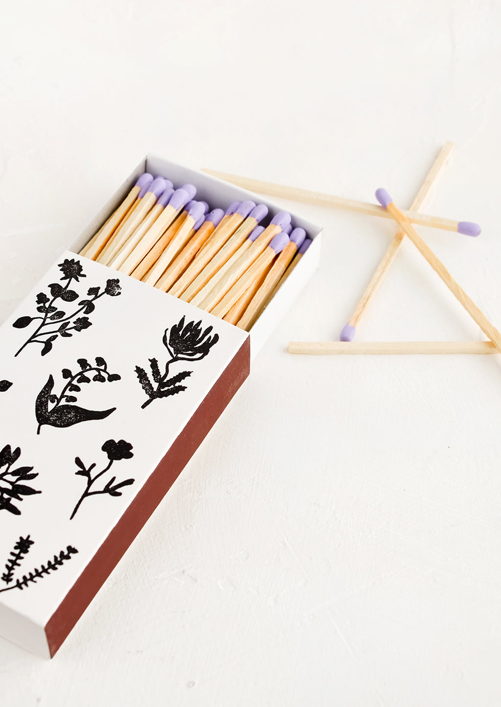 3: A matchbox in white with black botanical print, housing purple tipped matches.