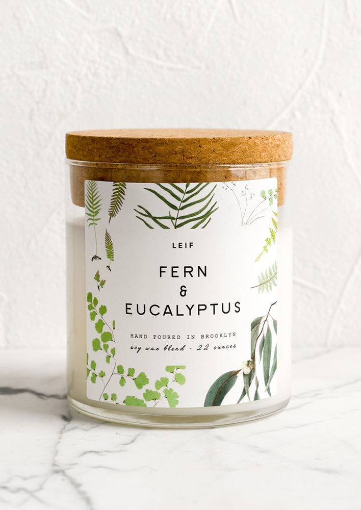 A glass candle with a cork lid and white botanical printed label reading "fern and eucalyptus".