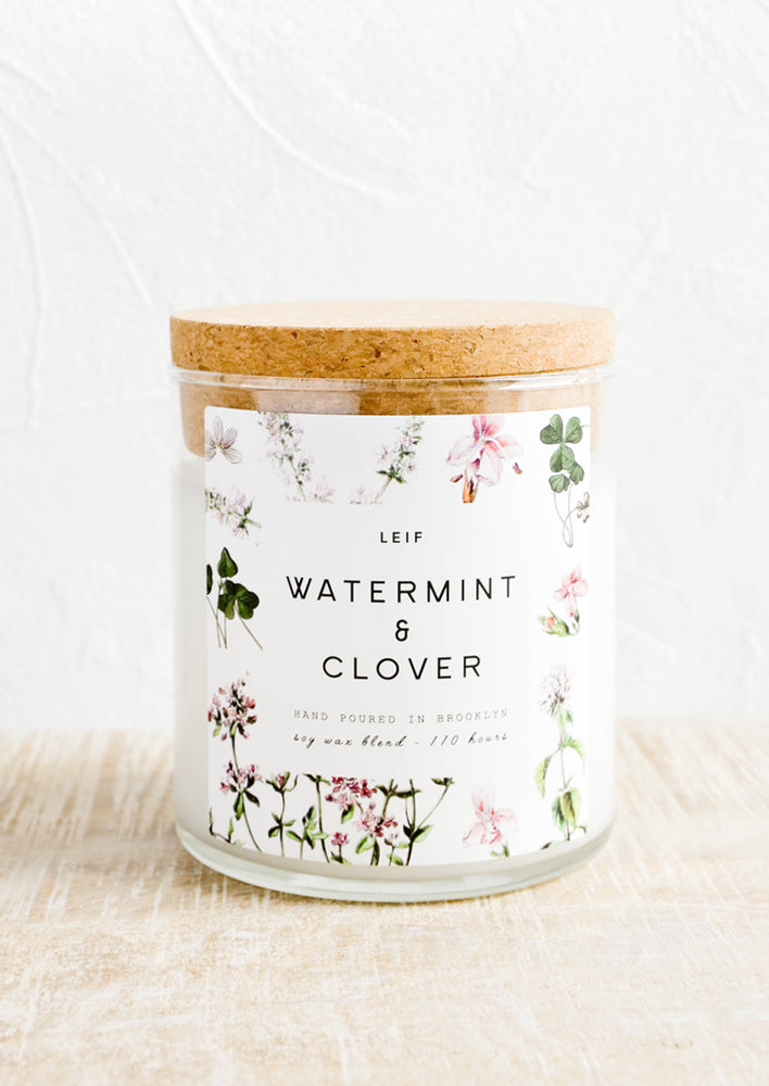 A glass candle with a cork lid and white botanical printed label reading "watermint and clover".
