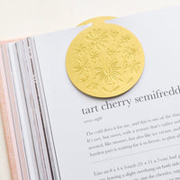 2: A circular brass bookmark with etched botanical pattern, marking a recipe page.