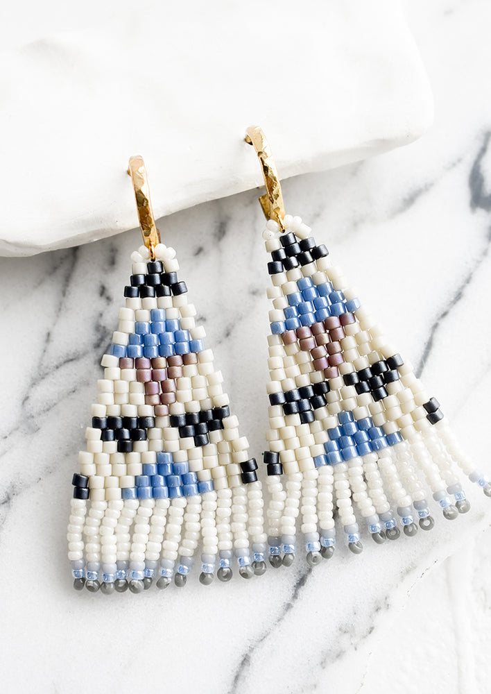 A pair of triangular shaped beaded earrings with geometric pattern in blue and cream.