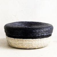 Carbon: A shallow, round storage basket with lid, woven from dried palm leaf in two-tone design.