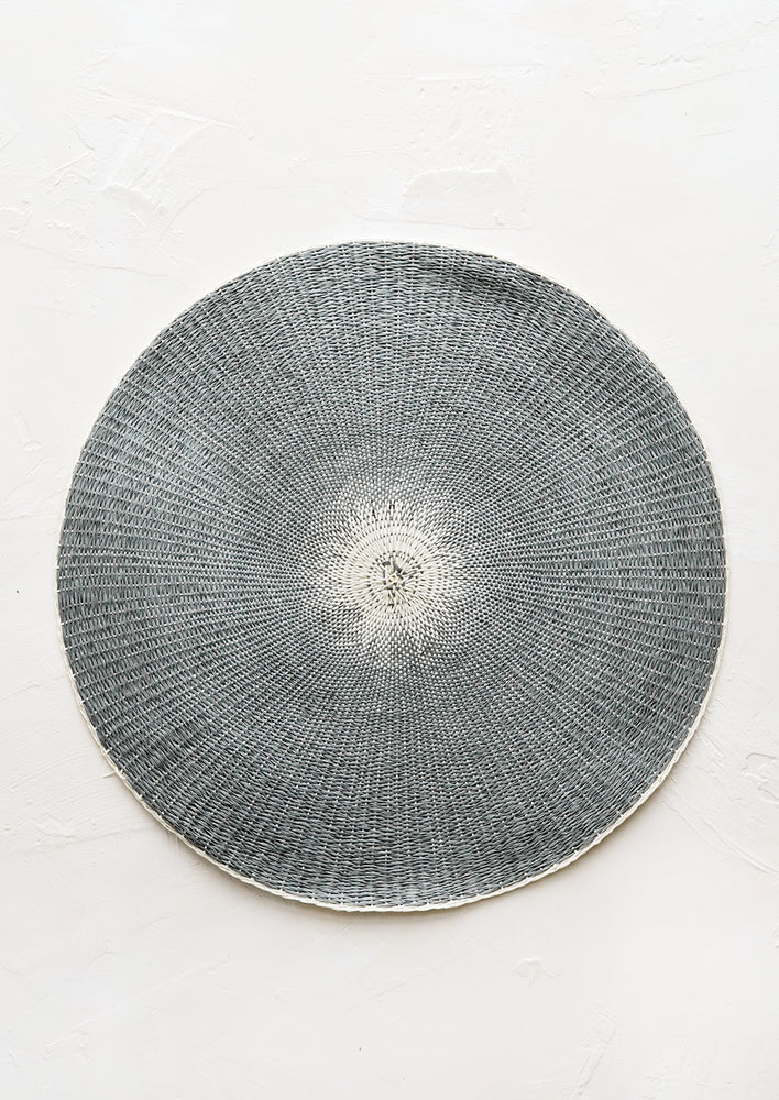 A round straw placemat in dark grey with white spot at center.