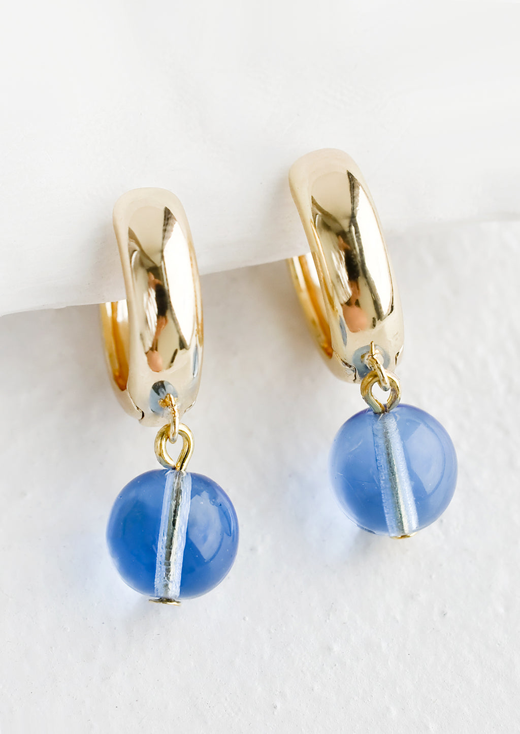 Sky Blue: A pair of gold huggie hoop earrings with single spherical blue glass bead detail.