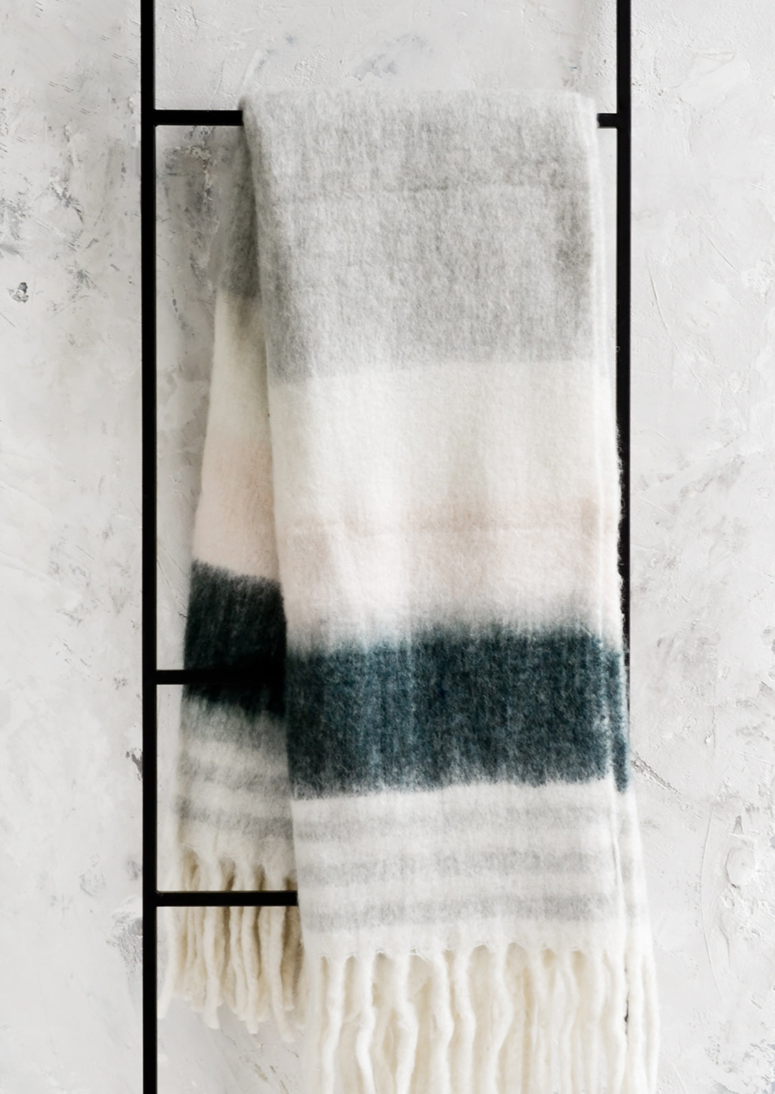 A striped throw blanket in pink and grey with fringe trim on a black ladder.