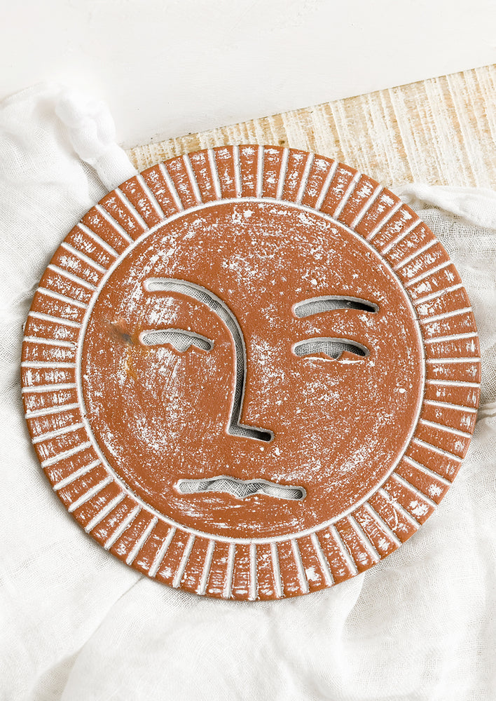 A rust colored cast iron trivet in the shape of a sun with cutout face detailing.