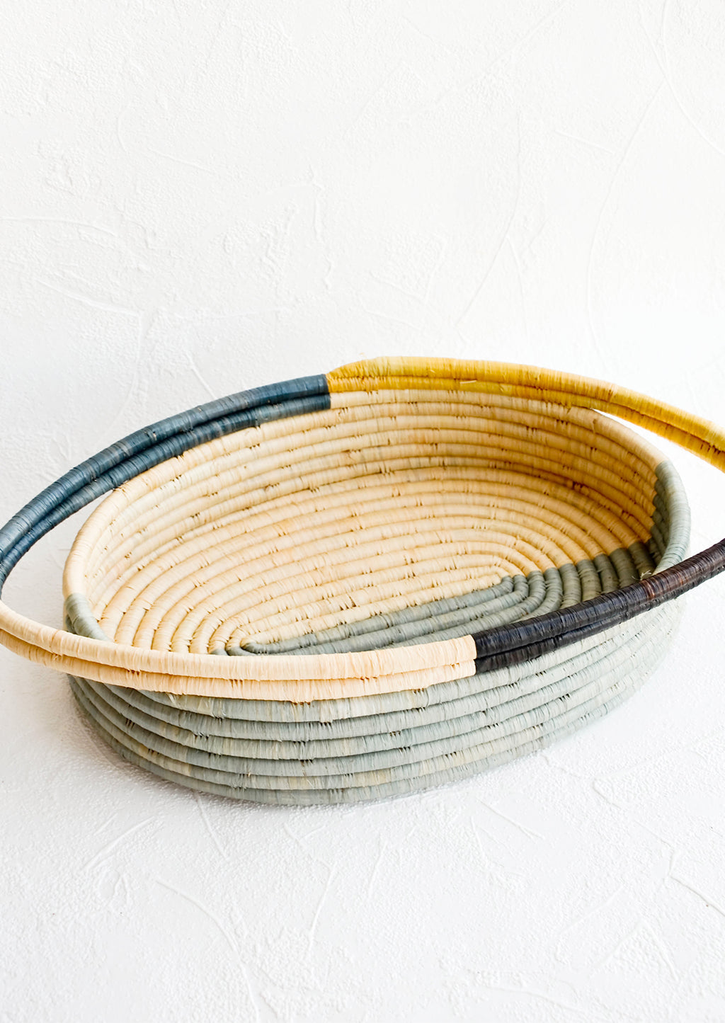 1: Oval bread basket in woven raffia with protruding handles at either side. Blue and yellow colorblock pattern.