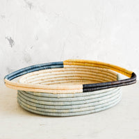 2: Oval bread basket in woven raffia with protruding handles at either side, sitting on a table.