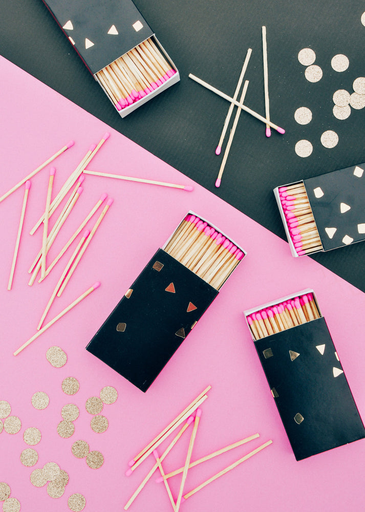 3: Three black and gold matchboxes are displayed amongst messy piles of pink-tipped matches on a black and pink surface.