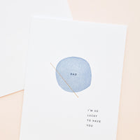 1: White greeting card with letter press printed blue circle at center reading "Dad". Bottom corner reads "I'm So Lucky To Have You".