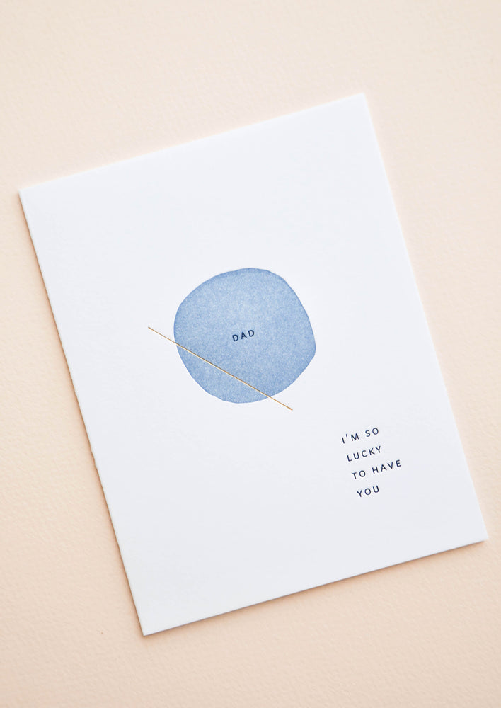 White greeting card with letter press printed blue circle at center reading "Dad". Bottom corner reads "I'm So Lucky To Have You".