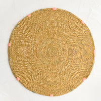 Tan: A round seagrass placemat in natural with grey and pink dashes embroidered around rim.