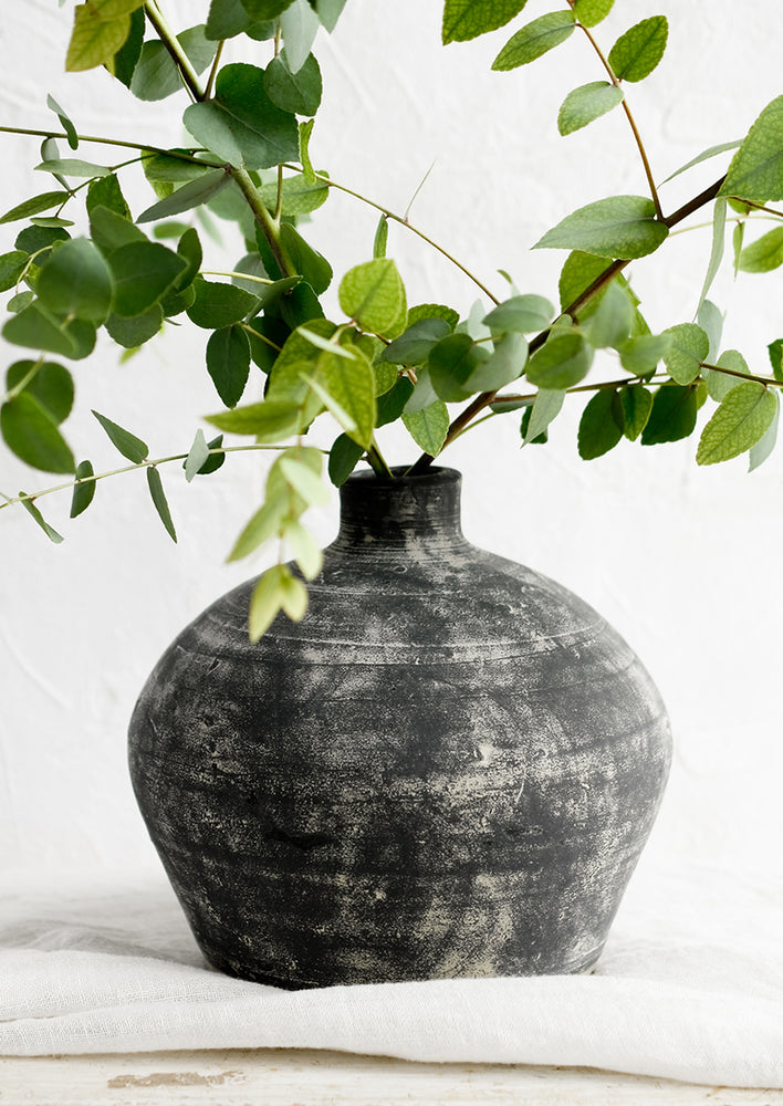 1: A distressed black clay vase with narrow opening, with eucalyptus branches.