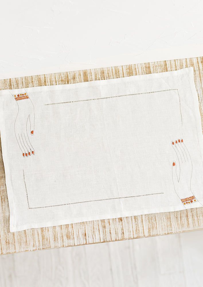 A white linen placemat with hemstitch border and embroidered hands at two corners.