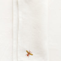1: A white linen napkin with single bee embroidery at corner in peach and mustard.
