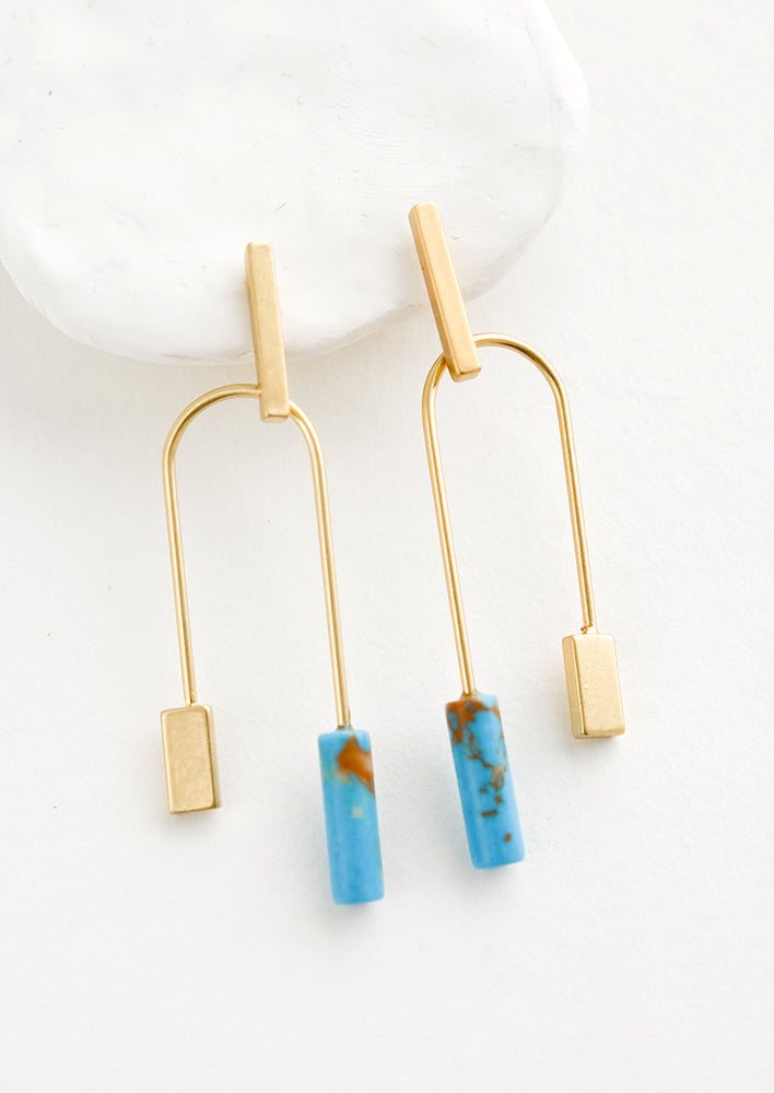 Gold / Turquoise: A pair of gold earrings with bar shaped top and arched bottom with turquoise gemstone on one side.