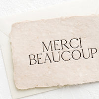 Single Card: A greeting card made from blush handmade paper and large letters spelling "Merci Beaucoup".