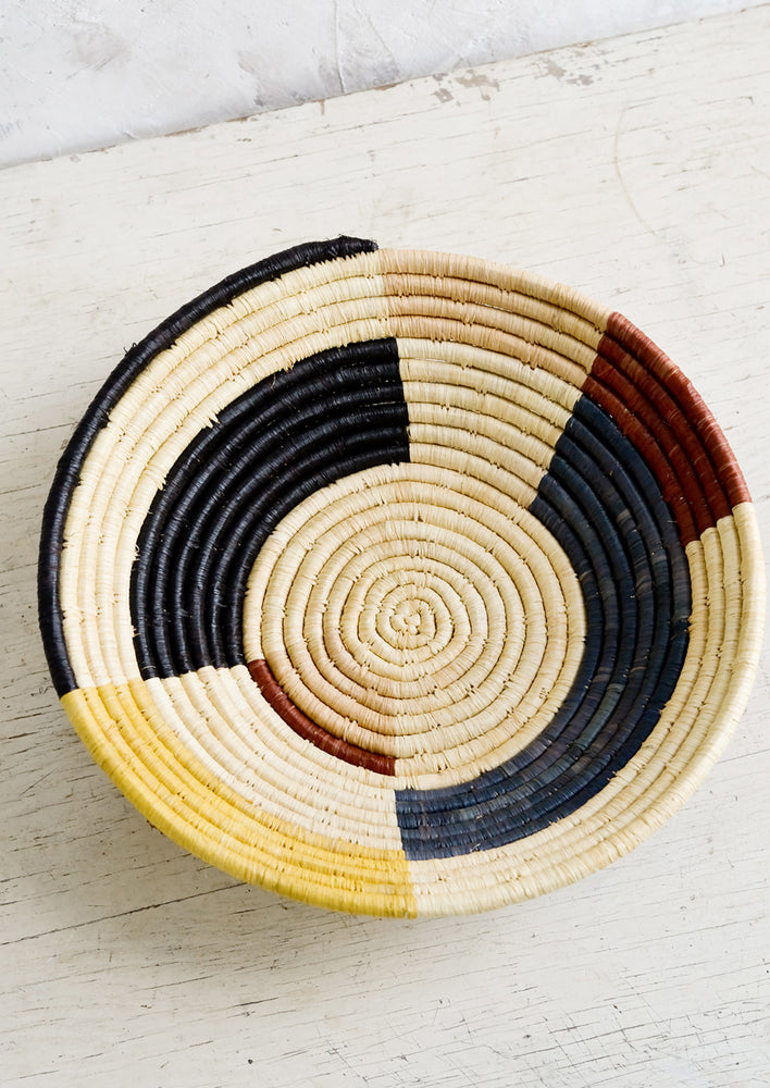 1: A round bowl made of raffia with a color blocked design in muted primary hues.