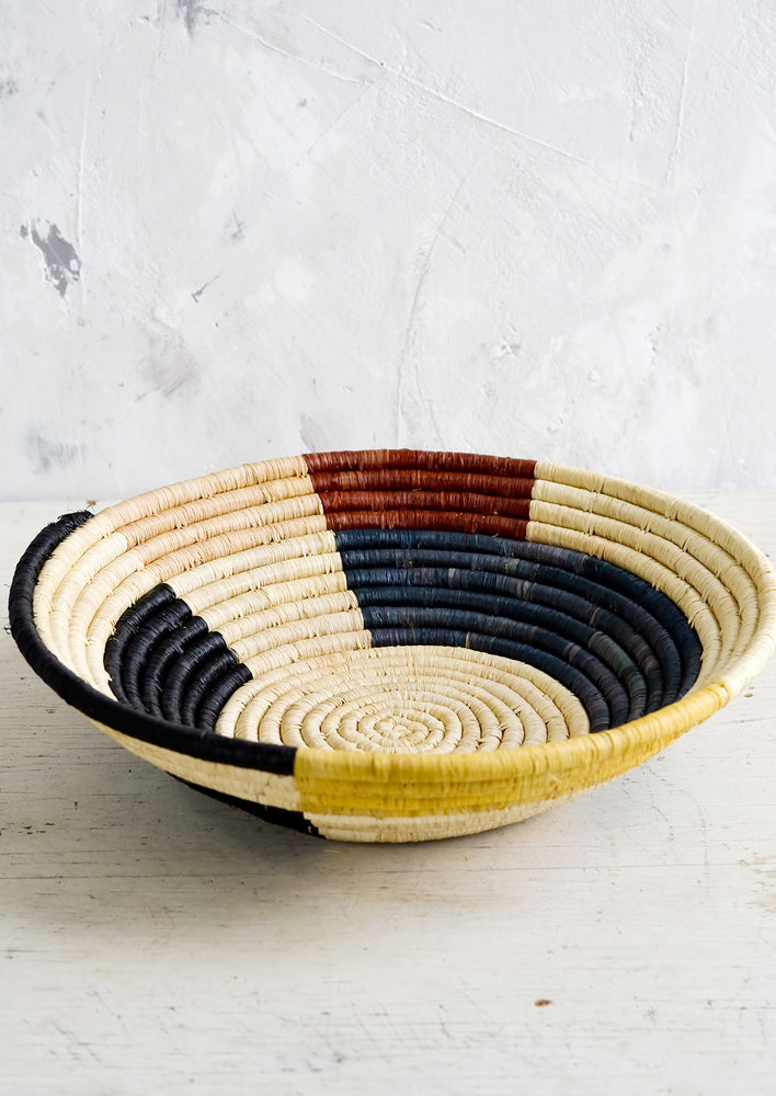 A round, shallow bowl made of raffia with a color blocked design in muted primary hues.