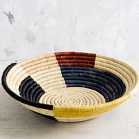 2: A round, shallow bowl made of raffia with a color blocked design in muted primary hues.