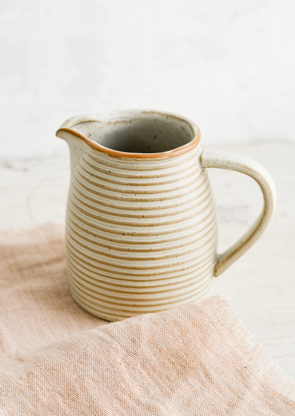 3: A tan ceramic pitcher with striped texture on top of pink linen.