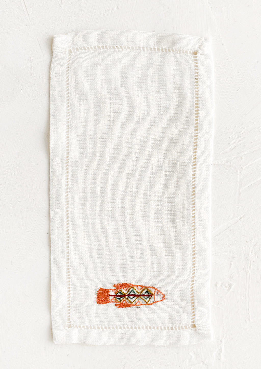 1: An white linen cocktail napkin with embroidered fish.