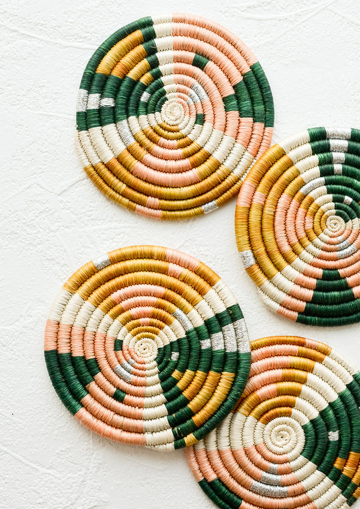 Four woven round sweetgrass coasters in pink, ochre, and dark green pattern with silver accents.
