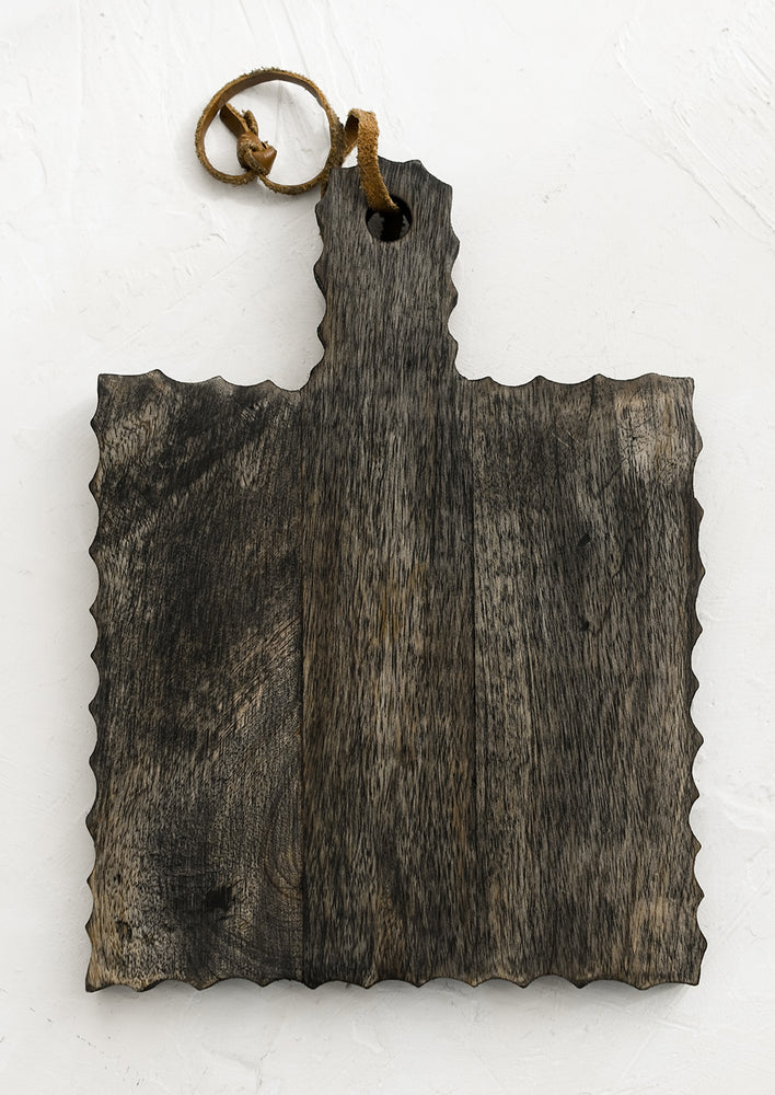 A square, paddle-shaped cutting board with carved/fluted edges and leather tie.