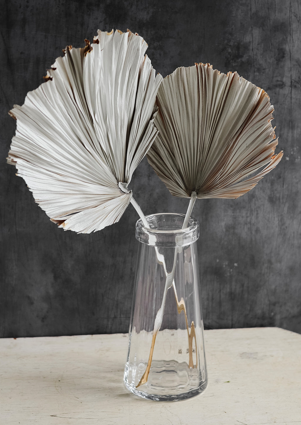 2: A tall, clear glass vase with two white dried fan palm leaves.