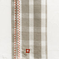 1: A plaid print napkin in beige and white with red embroidery detailing.