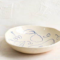 2: A round serving bowl in natural ceramic with blue fruit line drawings.