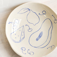 1: A round serving bowl in natural ceramic with blue fruit line drawings.