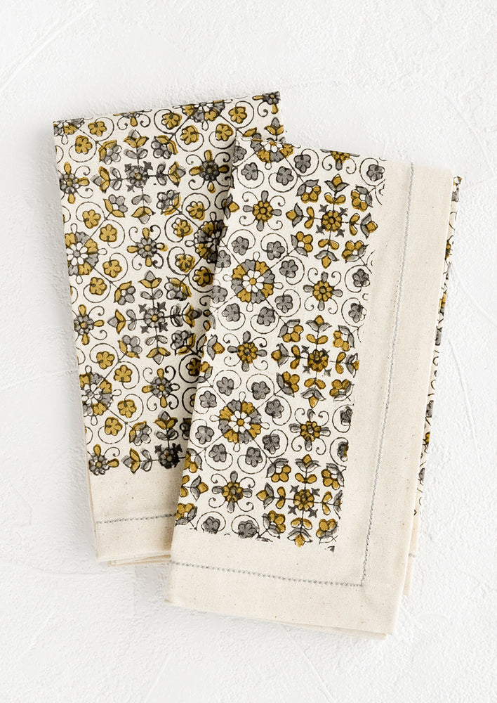 A pair of folded cotton napkins in floral print in grey and ochre.