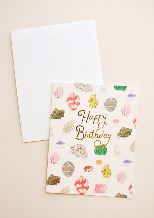 Notecard with colorful drawings of gems and minerals and the text "Happy Birthday" in gold foil, with white envelope.