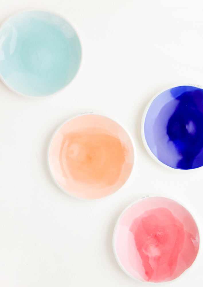 Four shallow porcelain dishes in light blue, dark blue, pale orange, and pink with shiny finishes and matte white rims.