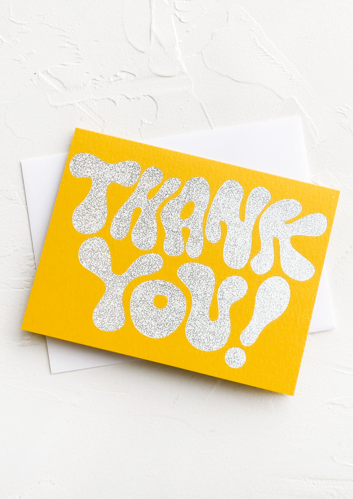 A yellow greeting card with silver glitter text reading "Thank you!"