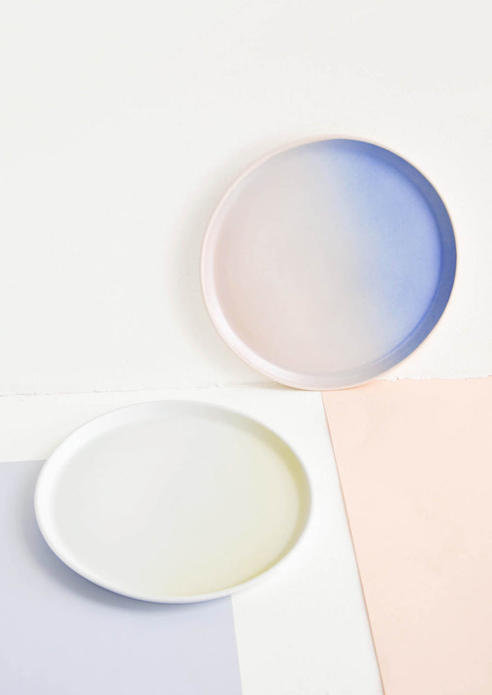 Two rimmed porcelain plates in blue and pink ombre and white and yellow ombre.