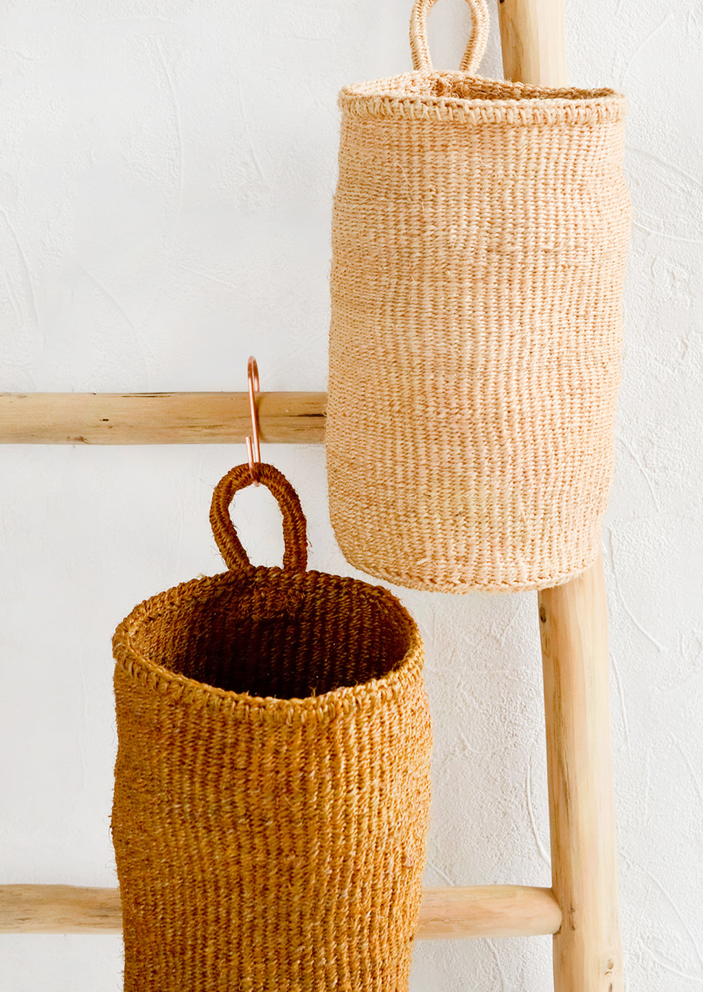 1: Two oblong cylindrical hanging baskets made from woven sisal, hanging on a display ladder.