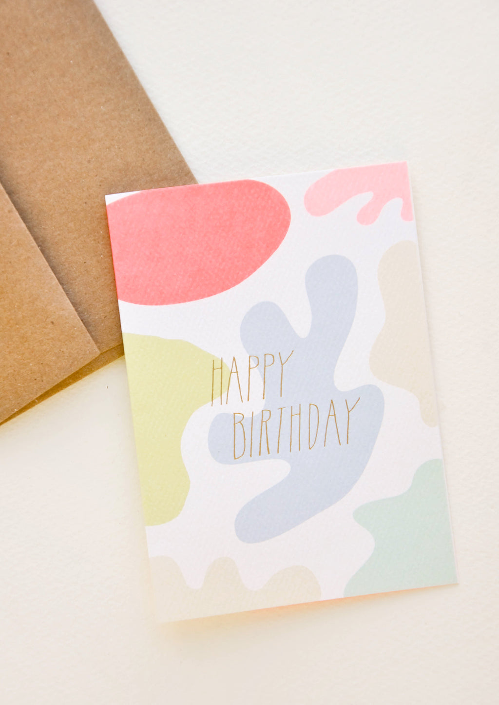 2: A brown paper envelope and greeting card patterned with differently colored blobs and the words "happy birthday" in uppercase gold foil.