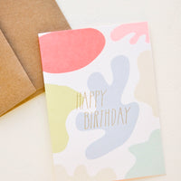 2: A brown paper envelope and greeting card patterned with differently colored blobs and the words "happy birthday" in uppercase gold foil.