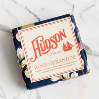 Rose Geranium: A square bar of soap in botanical printed packaging in Rose Geranium scent.