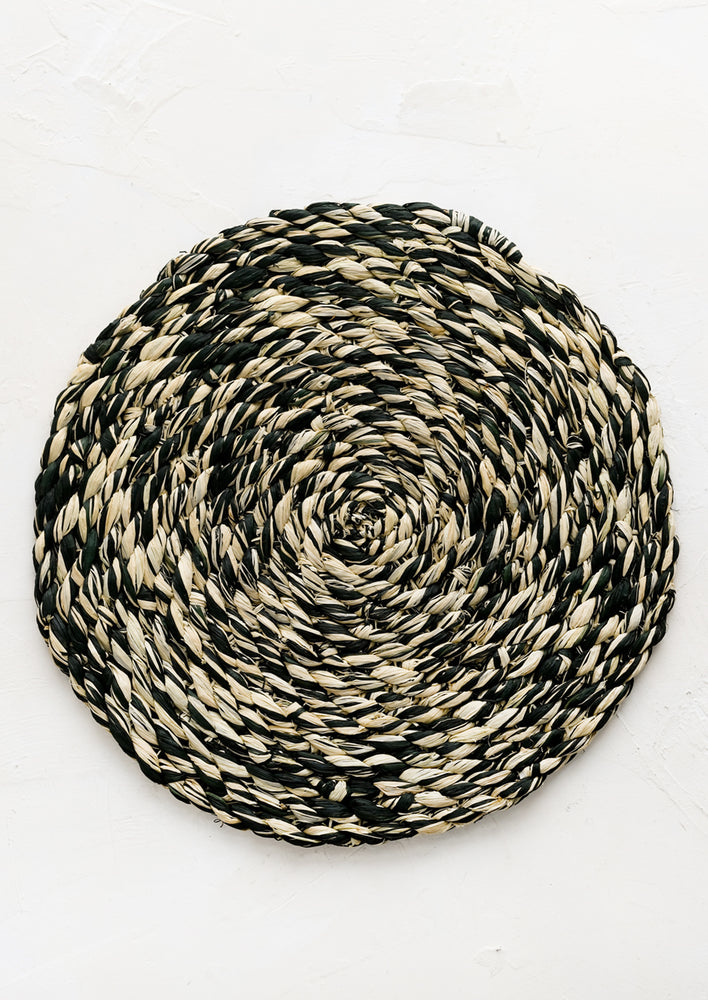 A round woven raffia placemat in natural and dark forest green.