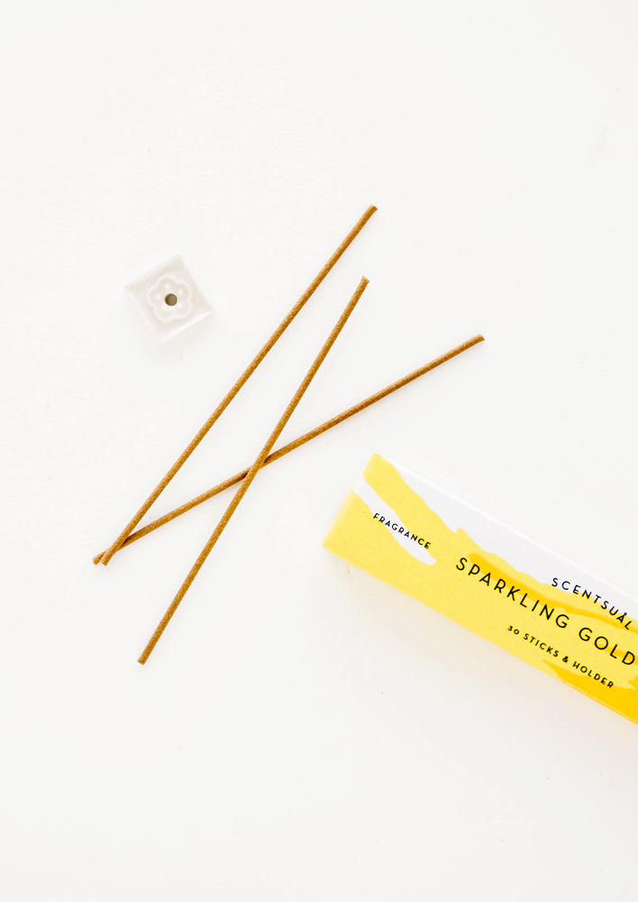 Three thin incense sticks laid out next to a small square holder and yellow cardboard box.
