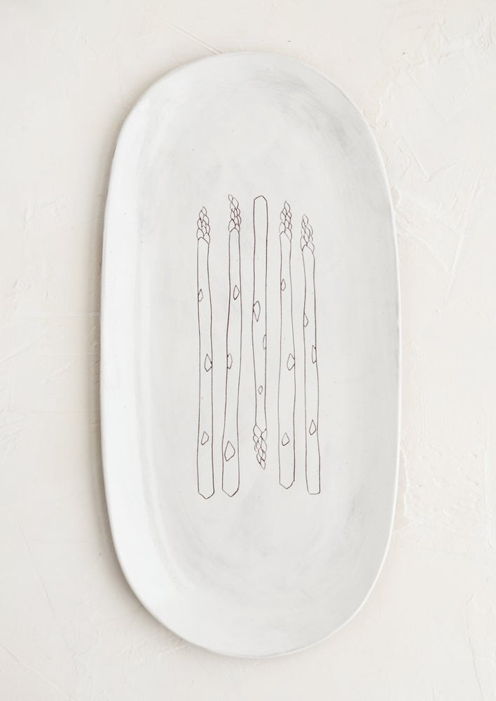 An oval shaped ceramic serving platter with hand drawn asparagus illustration at center.