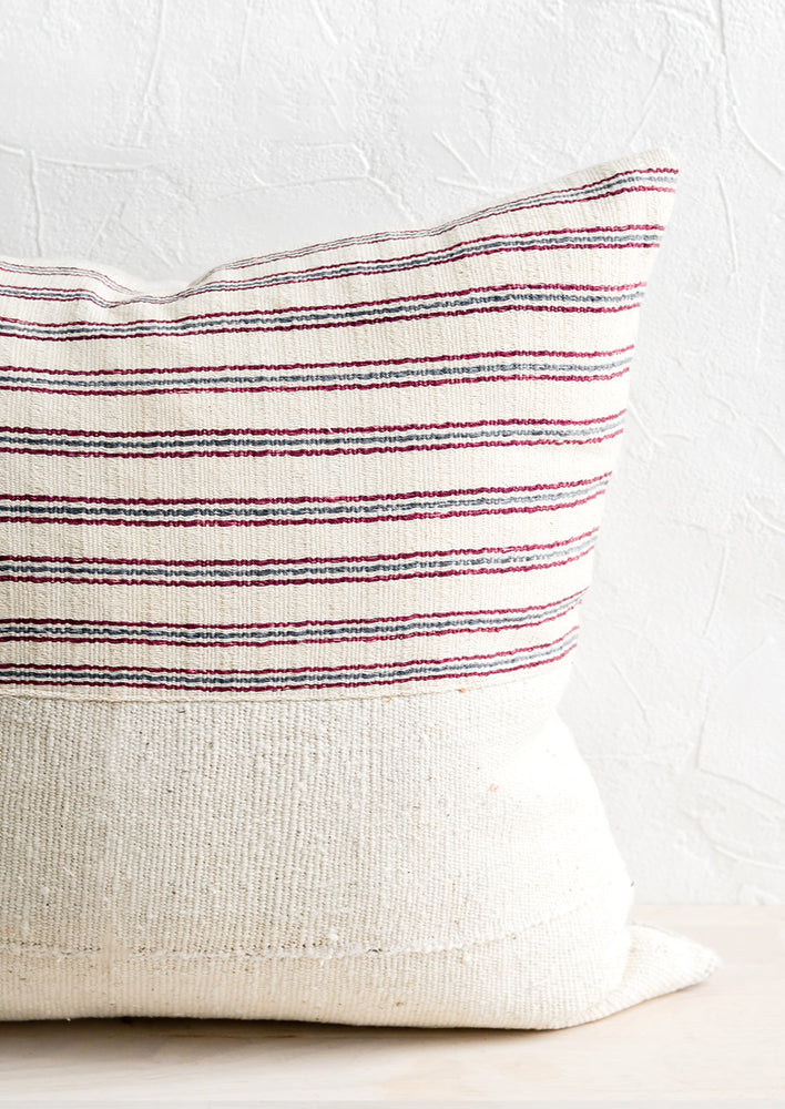 A throw pillow with top half in ivory, red and blue striped fabric and bottom half in natural mudcloth.