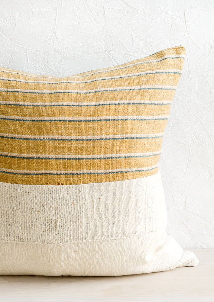 A throw pillow with top half in mustard, ivory & teal striped fabric and bottom half in natural mudcloth.