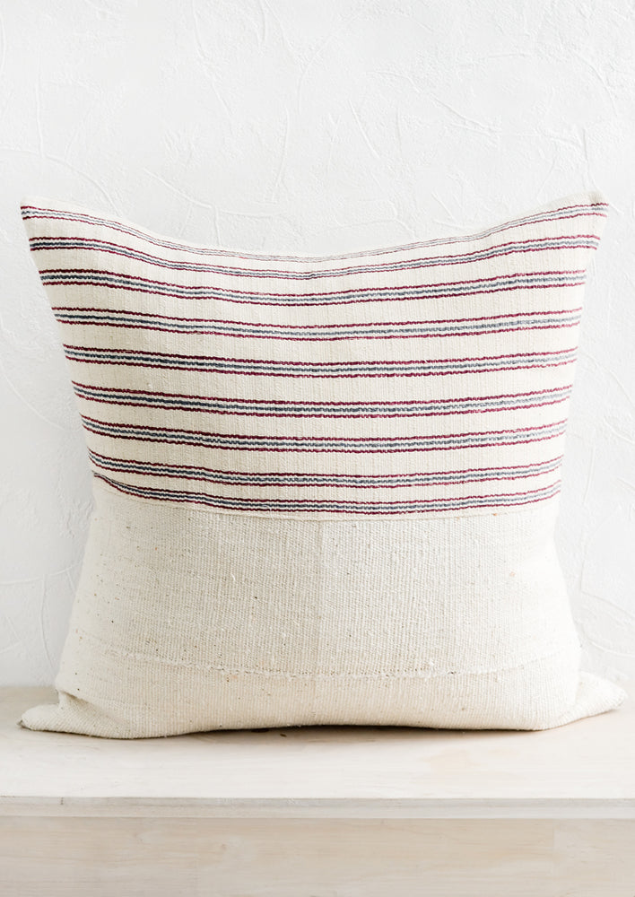 A throw pillow with top half in ivory, red and blue striped fabric and bottom half in natural mudcloth.