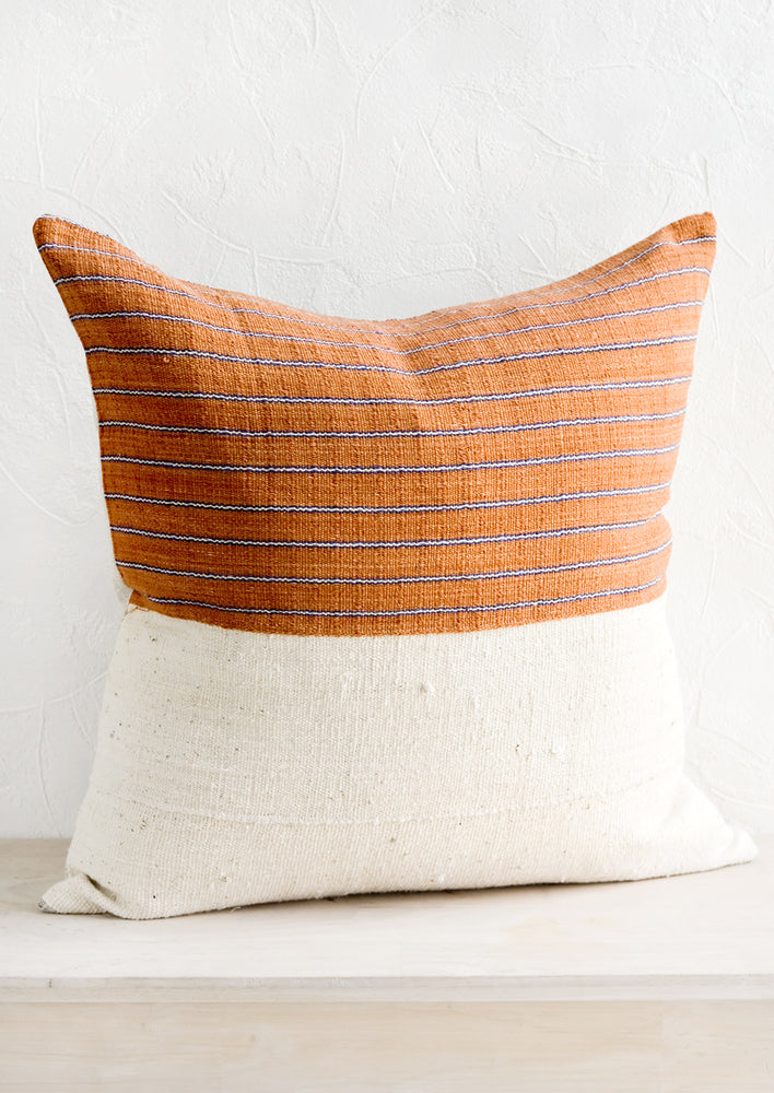 2: A throw pillow with top half in rust & indigo striped fabric and bottom half in natural mudcloth.