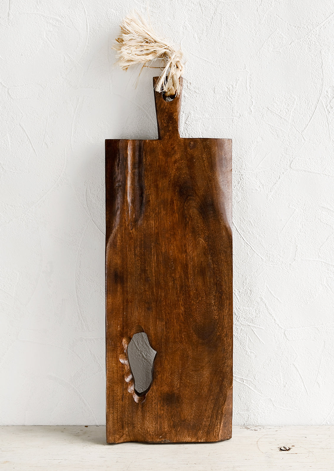 A rectangular wooden cutting board with knot cutout detail and twine strap handle.