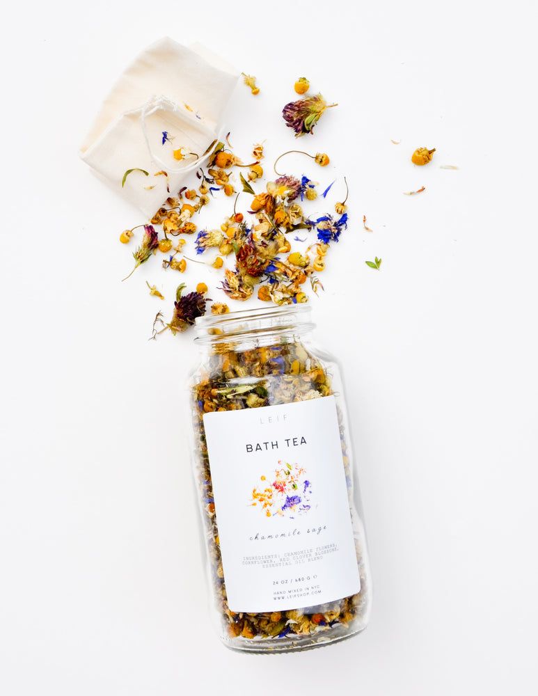 A mix of dried botanicals and a small muslin pouch spill out of a glass jar with a white label.